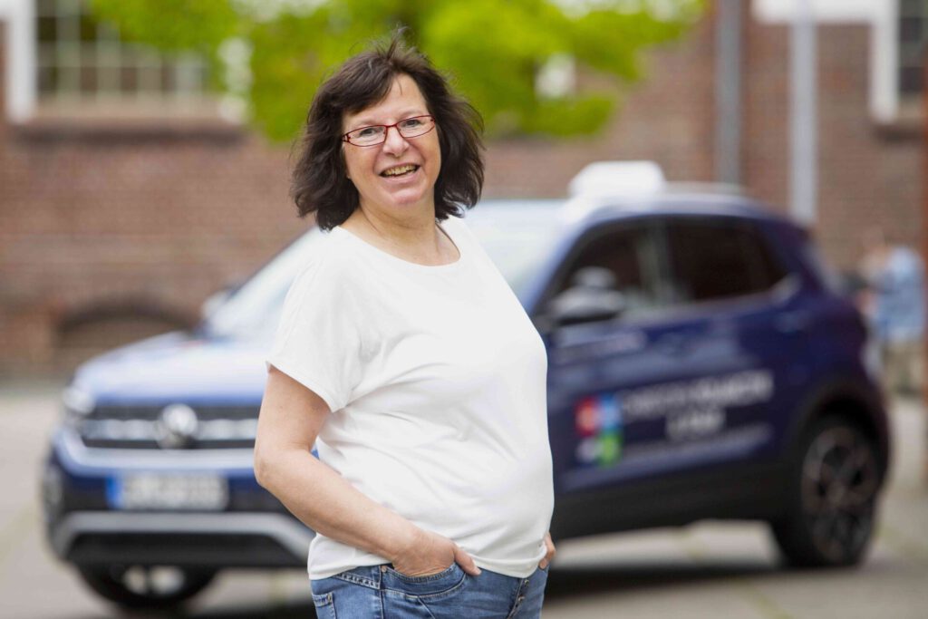 Eine Frau mit roter Brille und weißem T-Shirt steht vor einem Auto und lächelt, während das Hintergrundbild unscharf bleibt.