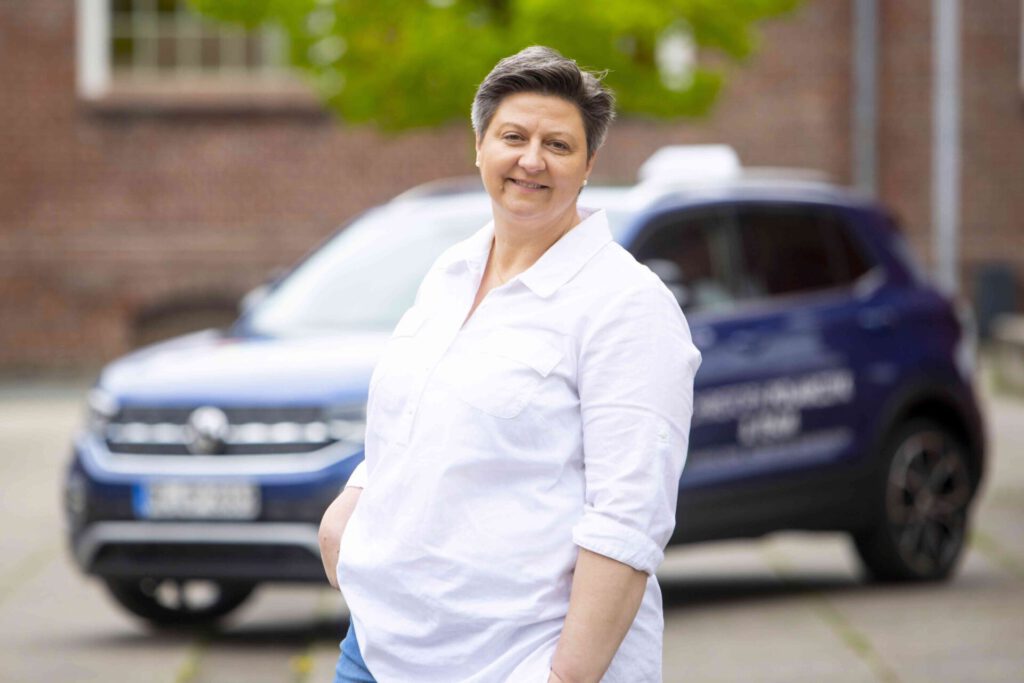 Freundliche Fahrlehrerin mit kurzhaarigem grauem Haar in weißem Hemd vor einem Pkw, im Hintergrund unscharfe Gebäudefassade und Grün.