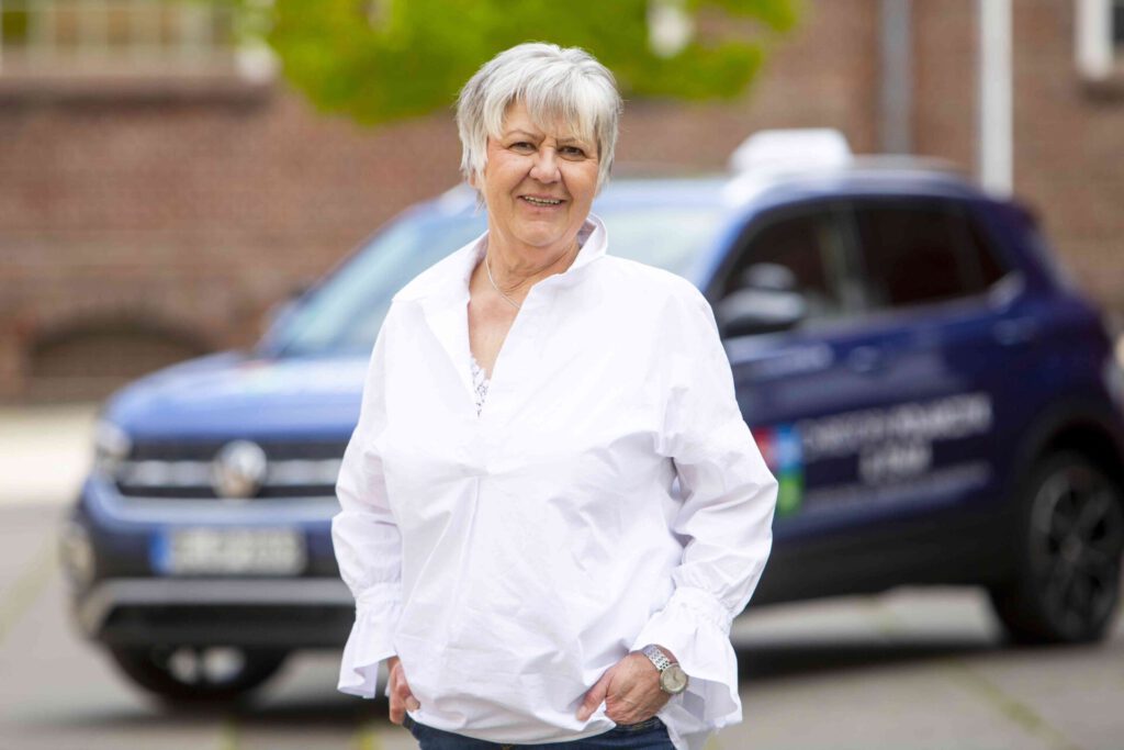 Lächelnde Fahrlehrerin mit kurzem grauem Haar und weißem Hemd steht neben einem Pkw im Freien, im Hintergrund unscharfe Gebäude.