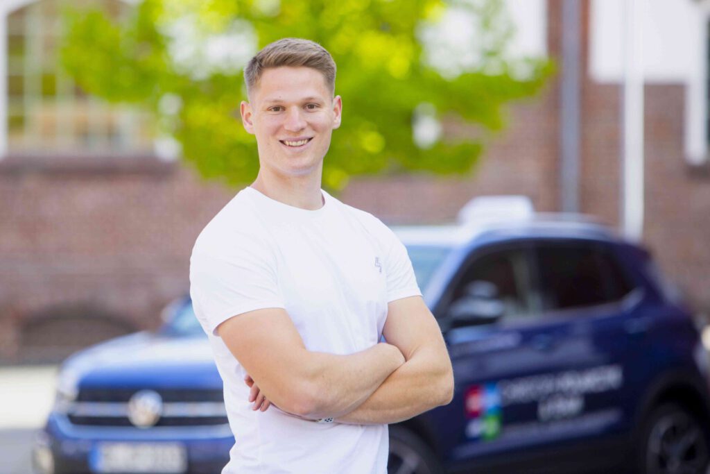 Lächelnder Fahrlehrer mit weißem T-Shirt und verschränkten Armen vor einem Auto, im Hintergrund sattgrünes Grün und Gebäude.