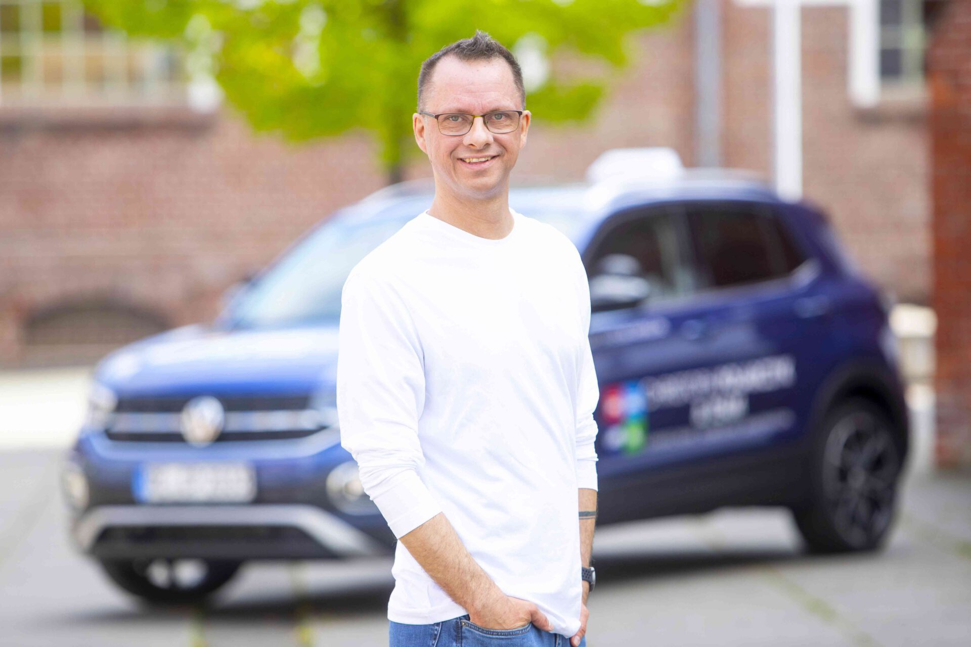 Freundlicher Fahrlehrer mit Brille im weißen Hemd vor einem blauen Auto, mit grünem Hintergrund und unscharfer Fassade im Hintergrund.