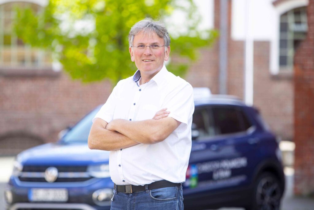 Grauhaariger Mann mit Brille steht mit verschränkten Armen vor einem blauen Auto, trägt ein weißes Hemd, lächelt im Freien.