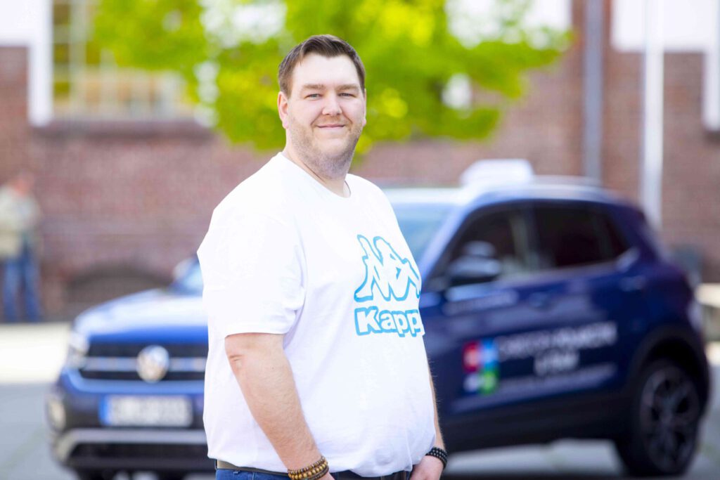 Ein Mann im weißen T-Shirt mit „Kapp“-Logo steht vor einem blauen Auto und blickt freundlich in die Kamera, im Hintergrund unscharf.