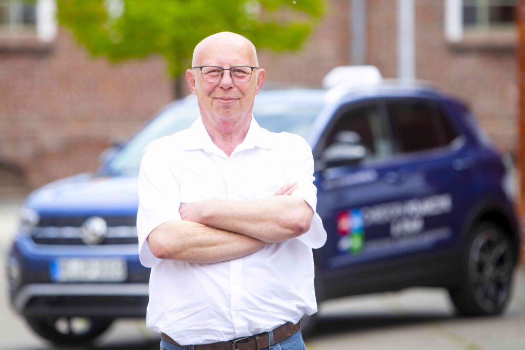 Mann mit Brille und weißem Hemd steht vor einem Auto und hat die Arme verschränkt, im Hintergrund ist ein Gartenbereich unscharf.