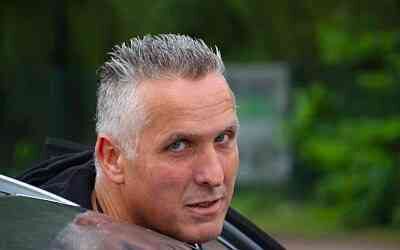 Graubärtiger Fahrlehrer mit kurzen grauen Haaren schaut aus einem Fahrzeug nach vorn, draußen vor Grün im Hintergrund bei Tageslicht.